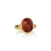A yellow gold ring featuring a large oval-cut red tourmaline center stone, accented by small dark purple tanzanite stones on the shoulders, set against a clean white background with a soft reflection.