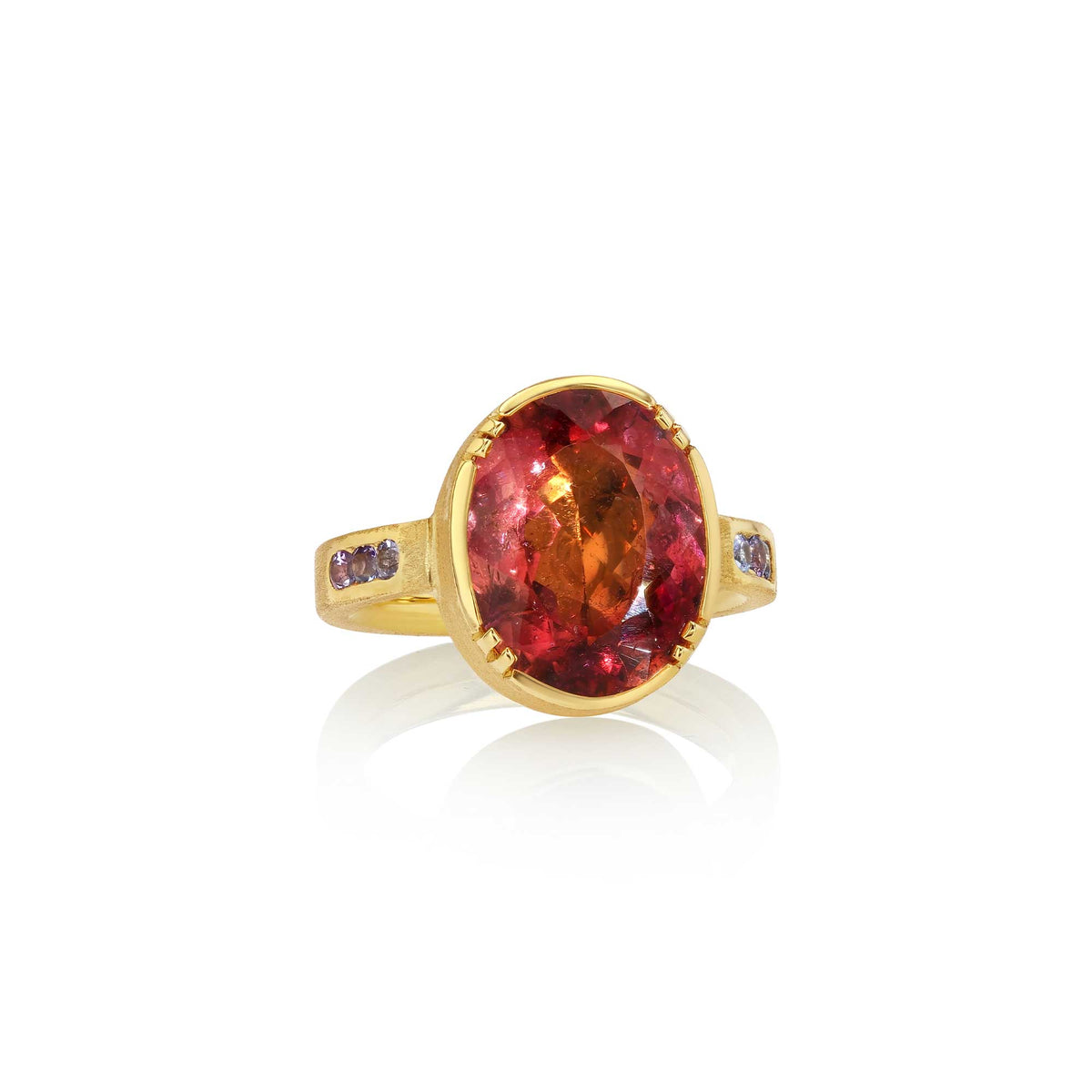 A yellow gold ring featuring a large oval-cut red tourmaline center stone, accented by small dark purple tanzanite stones on the shoulders, set against a clean white background with a soft reflection.