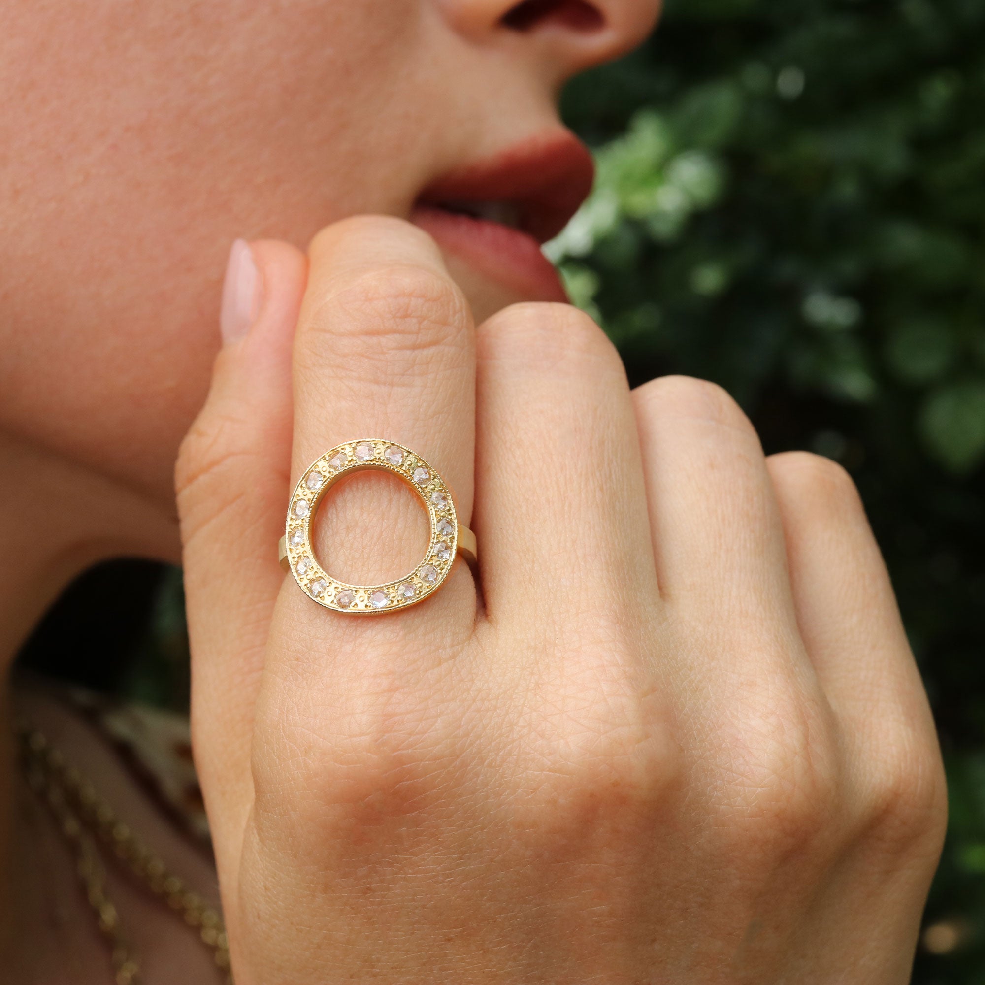 Close-up of a hand wearing a gold ring with daimond gemstones, against a blurred green background.