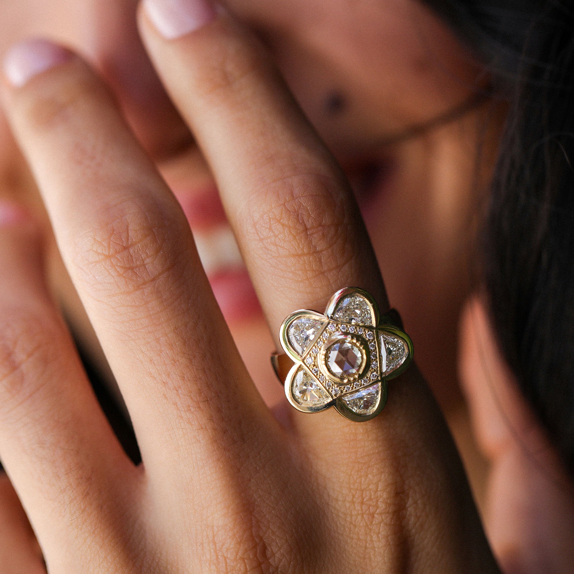 A gold flower-shaped ring with a central rosecut diamond gemstone and small diamond stones, worn on a person's finger.
