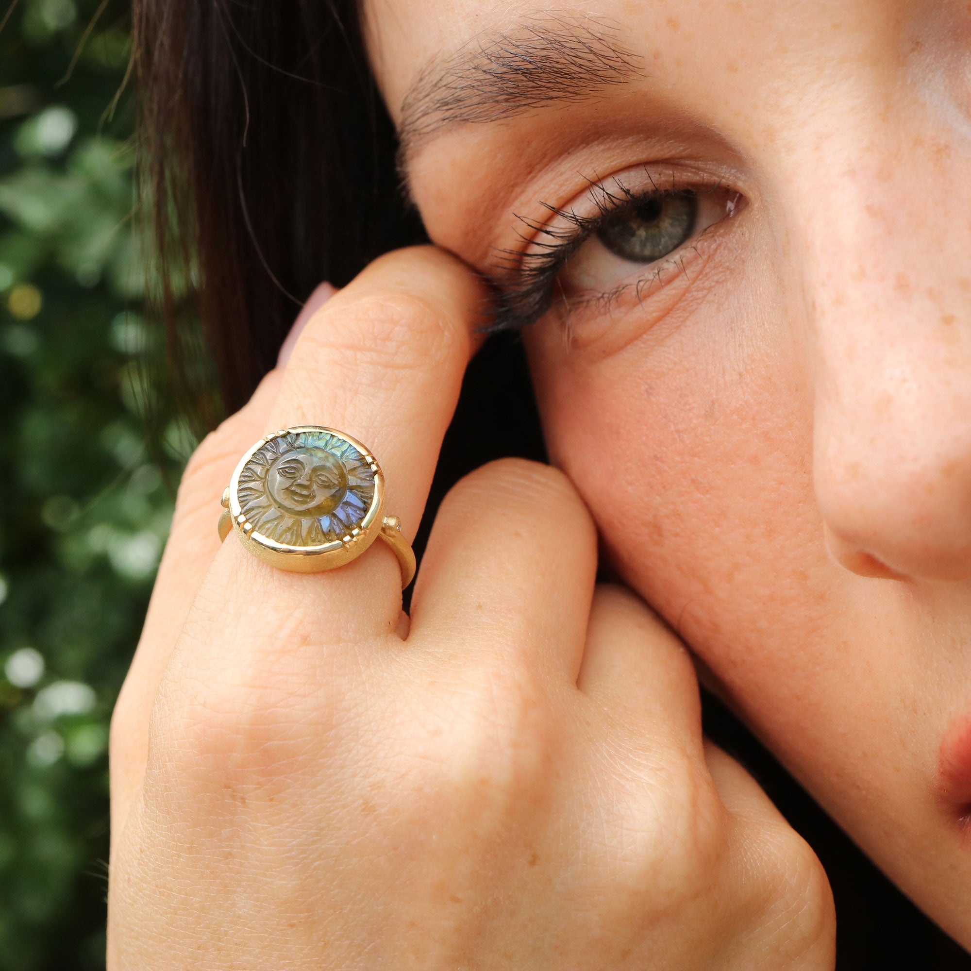 Close-up of a person wearing a ring with a labradorite sun face design, outdoors.