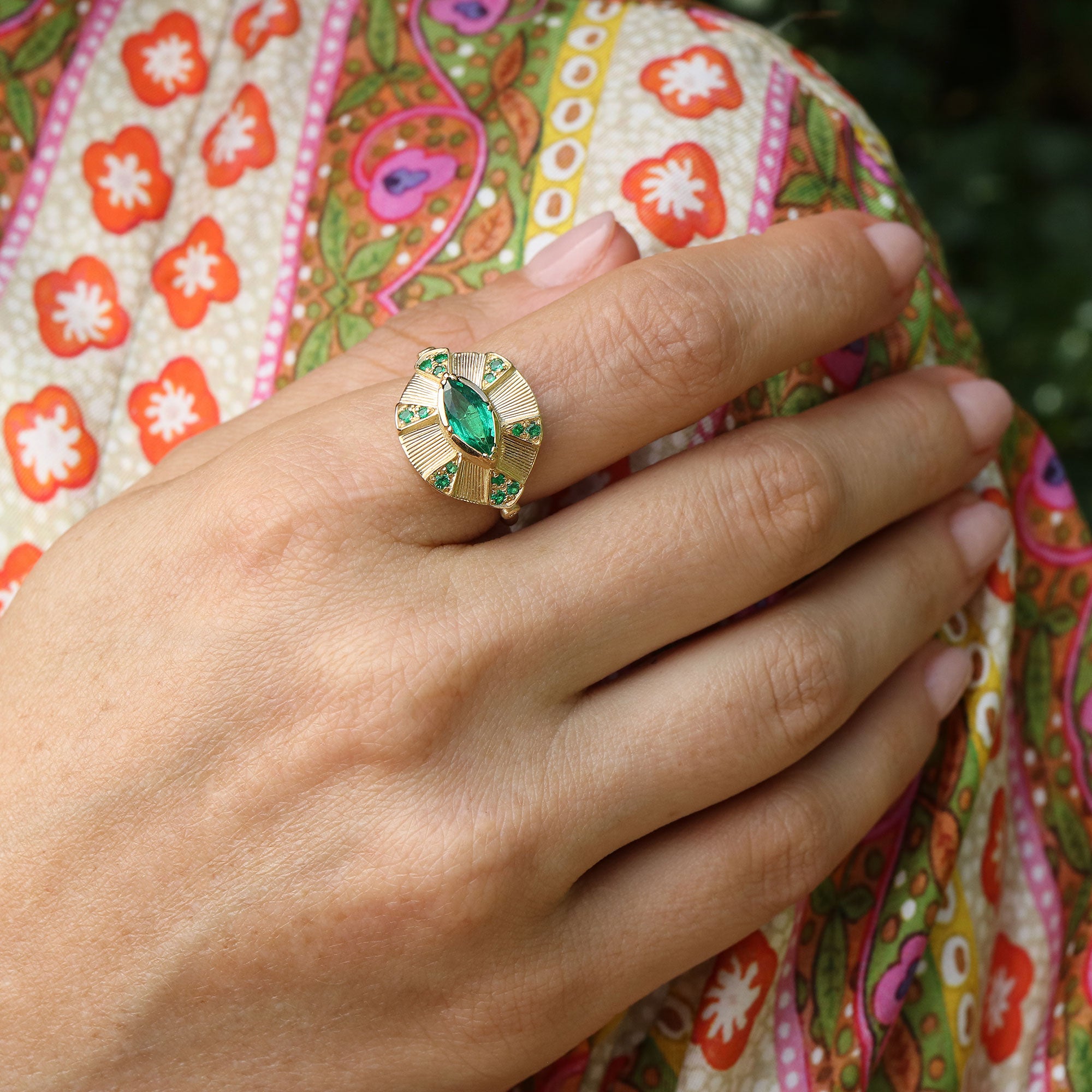 Gold ring with a green emerald gemstone on a hand against a colorful fabric background