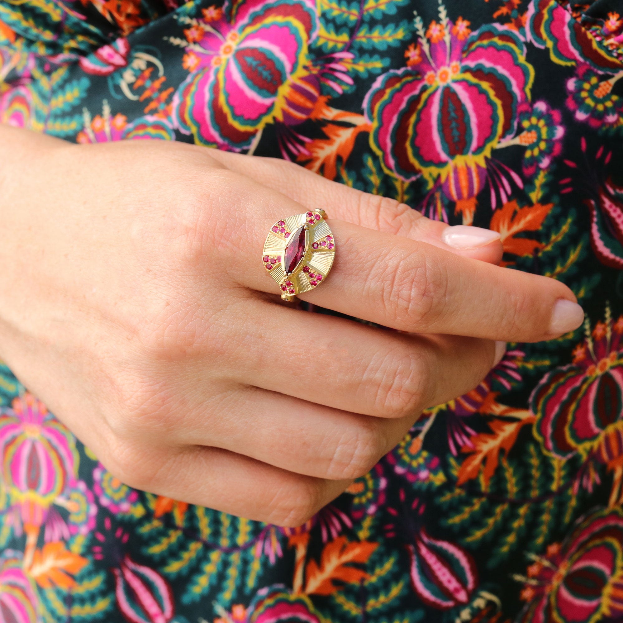 Gold ring with a red ruby gemstone on a hand against a colorful floral background