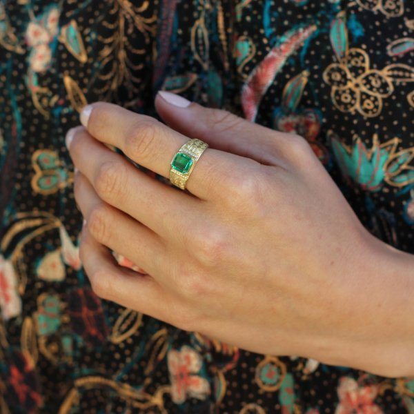 Hand wearing a gold ring with an emerald stone on a patterned fabric background