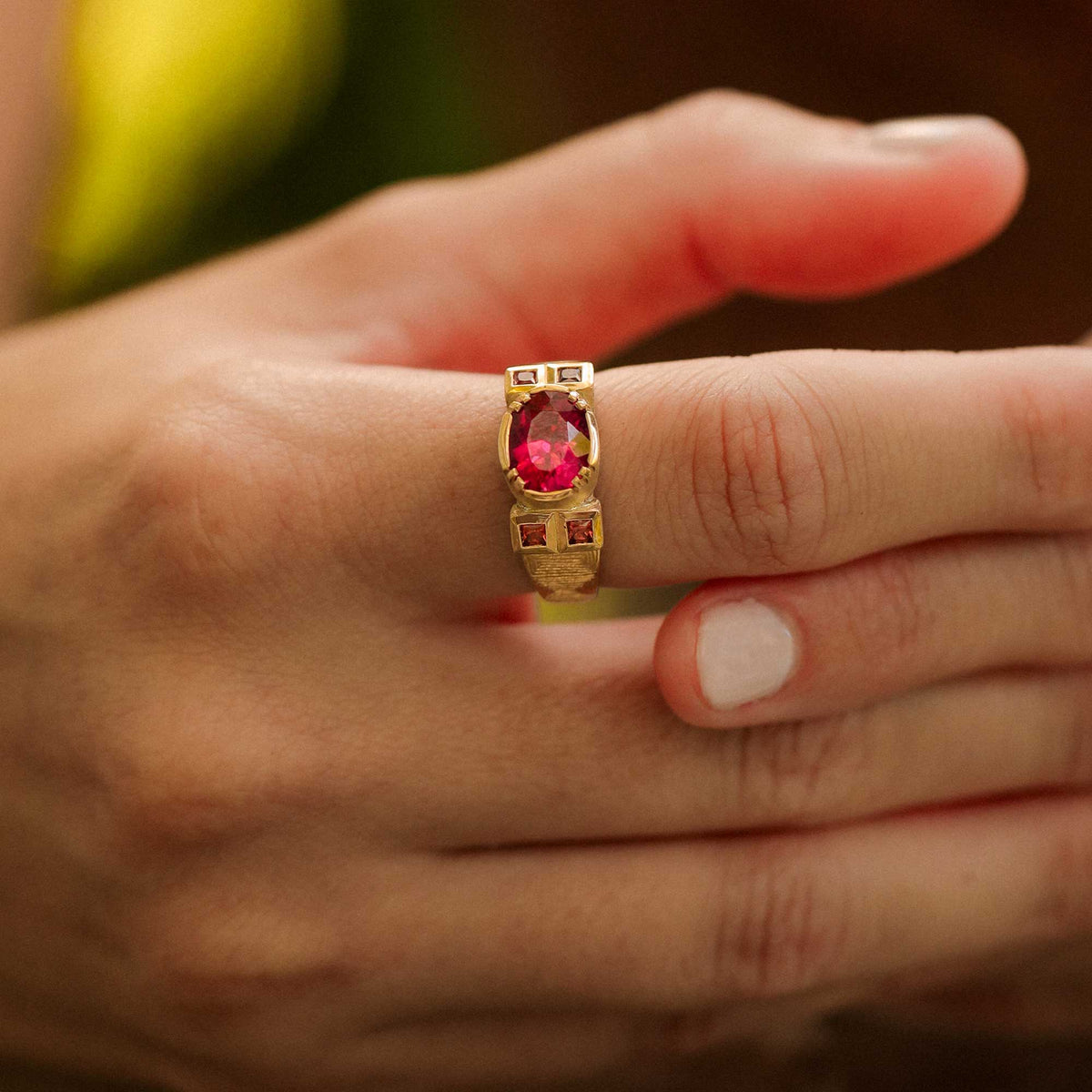An 18k yellow gold ring with a large oval pink tourmaline gemstone and smaller square pink sapphire stones on the sides with hand engraved detailing on either sides, worn on a person's finger.