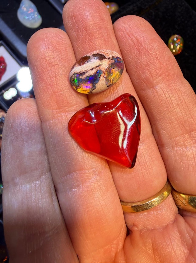 A close-up of a hand holding two polished stones: a heart-shaped red fire opal gemstone and an irregular opal stone with multicolored play-of-color patches. The hand also wears gold rings, and a dark jewelry display is blurred in the background.