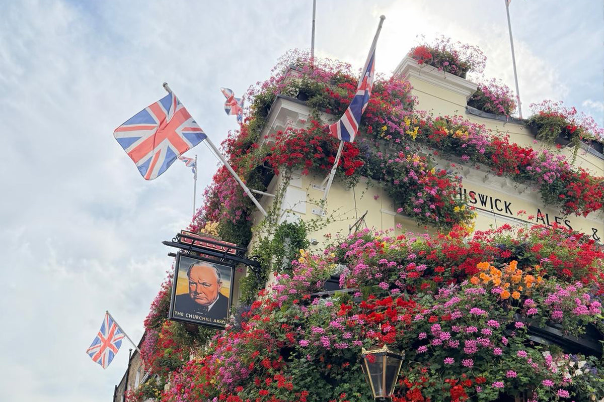 A white pub building covered in dense pink, red, and orange flowers, with Union Jack flags flying from the exterior and a hanging sign featuring a portrait. The sky is cloudy in the background.