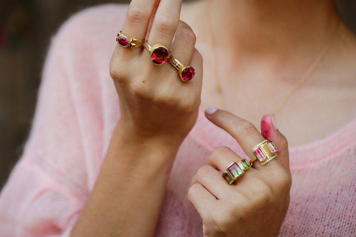 A close-up of a person wearing multiple gold rings with colorful tourmaline gemstones, including pink, green, and purple stones, while wearing a pink knit top and eating a pink macaron. Jewelry by Brooke Gregson.